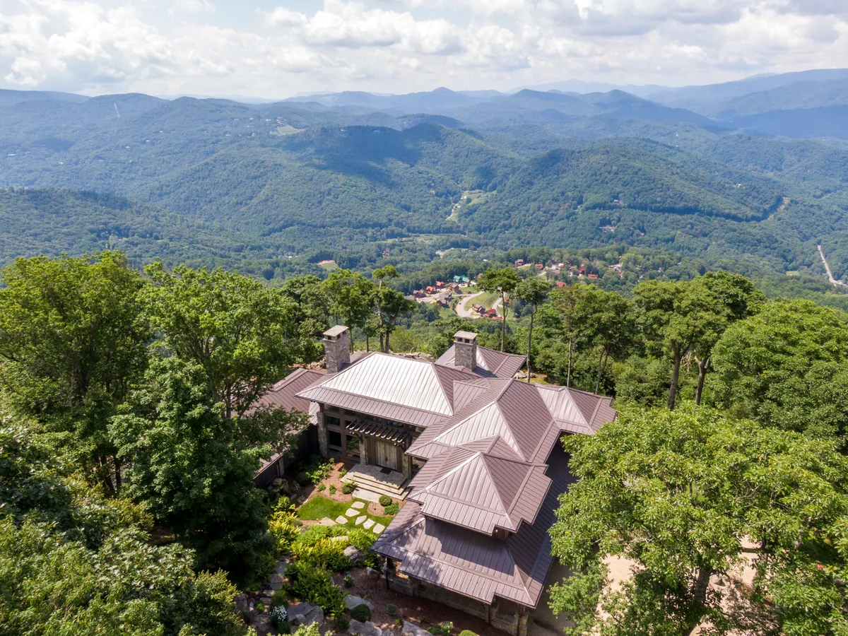 An elegant mountain home framed by lush trees, with expansive windows and a welcoming patio. The soft morning light enhances the house's stone and wood features, while the surrounding mountains hint at breathtaking sunrise and sunset views. The outdoor area feels private and tranquil, surrounded by natural beauty.