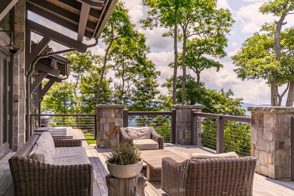 A tranquil deck scene featuring wicker seating, a wooden deck, stone pillars, lush greenery, and a sweeping mountain view. The area is perfect for both quiet reflection and lively group gatherings, immersed in natural beauty.