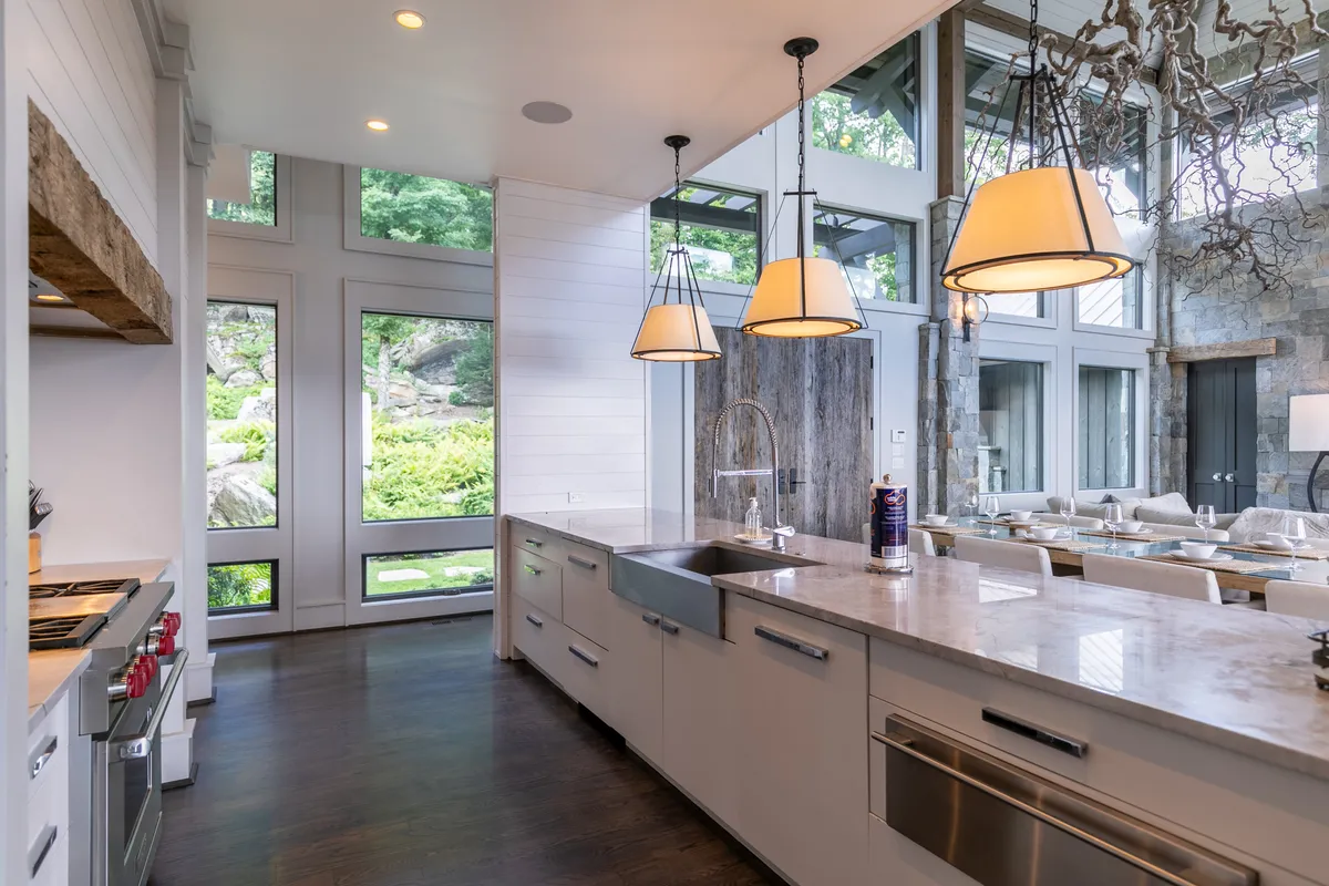 A welcoming, modern kitchen with a large island sits at the heart of the home. Hanging light fixtures illuminate the space, while expansive windows provide a view of the outdoor landscape. The kitchen is outfitted with high-end appliances and flows seamlessly into an open-plan layout ideal for gatherings.