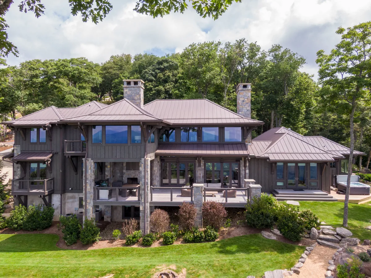 This exterior image highlights the home's inviting stone pillars, large windows, metal roof, and landscaped garden, with a patio area ideal for gatherings and relaxation. The natural setting and expansive deck showcase the transition from indoor comfort to outdoor adventure and seasonal enjoyment.