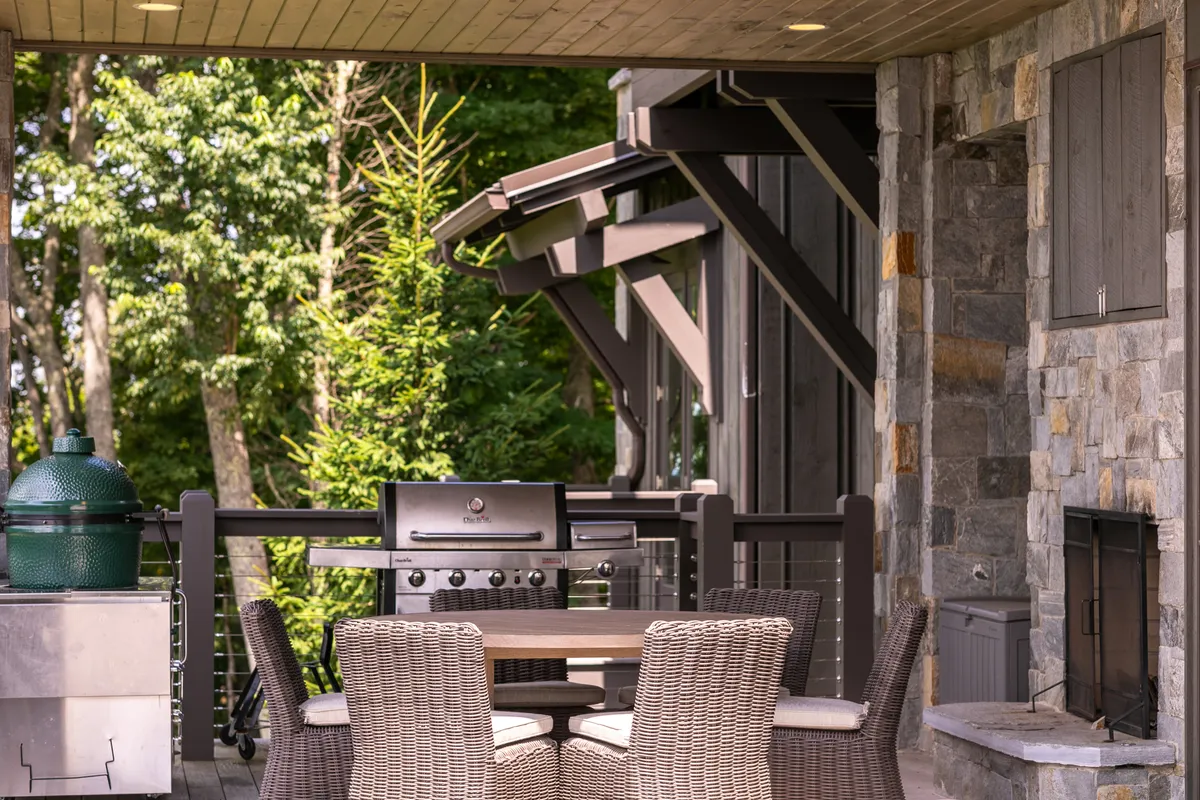 A spacious outdoor dining and lounge area featuring a grill, dining table, comfortable seating, and a fireplace surrounded by lush greenery. The ambiance is welcoming and communal, ideal for gatherings and alfresco meals.