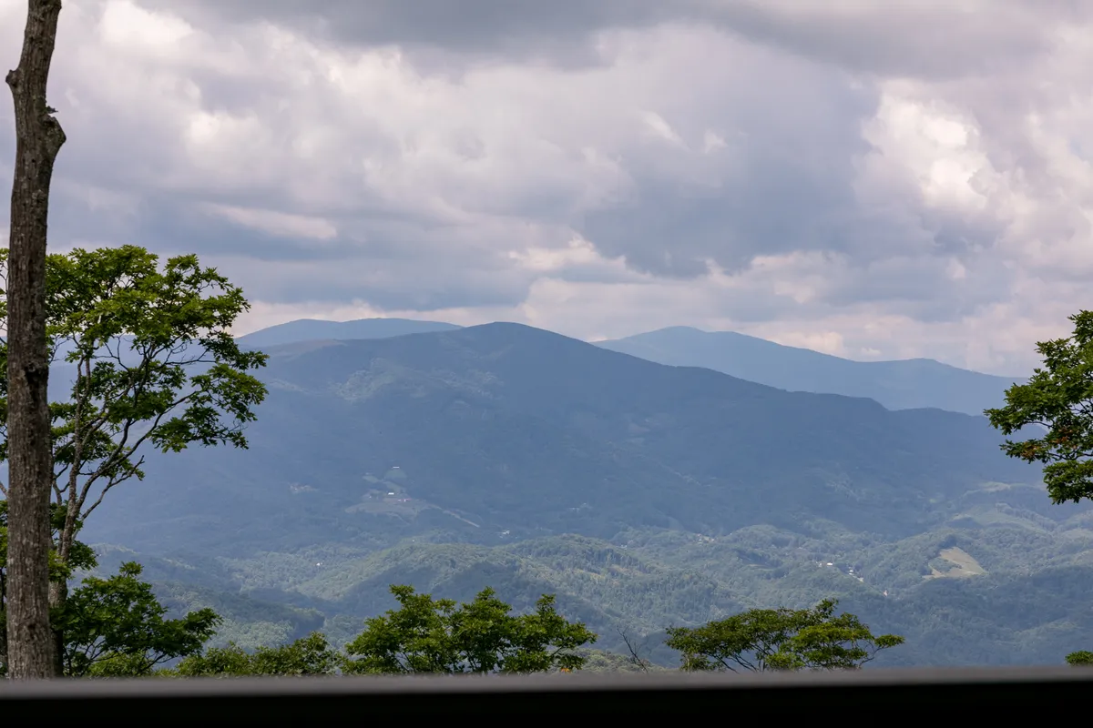 A sweeping mountain view with a dramatic range stretching across the horizon, under a tranquil sky with some clouds, framed by trees. The landscape is inviting and hints at scenic hiking, mountain biking, and opportunities for nature photography.