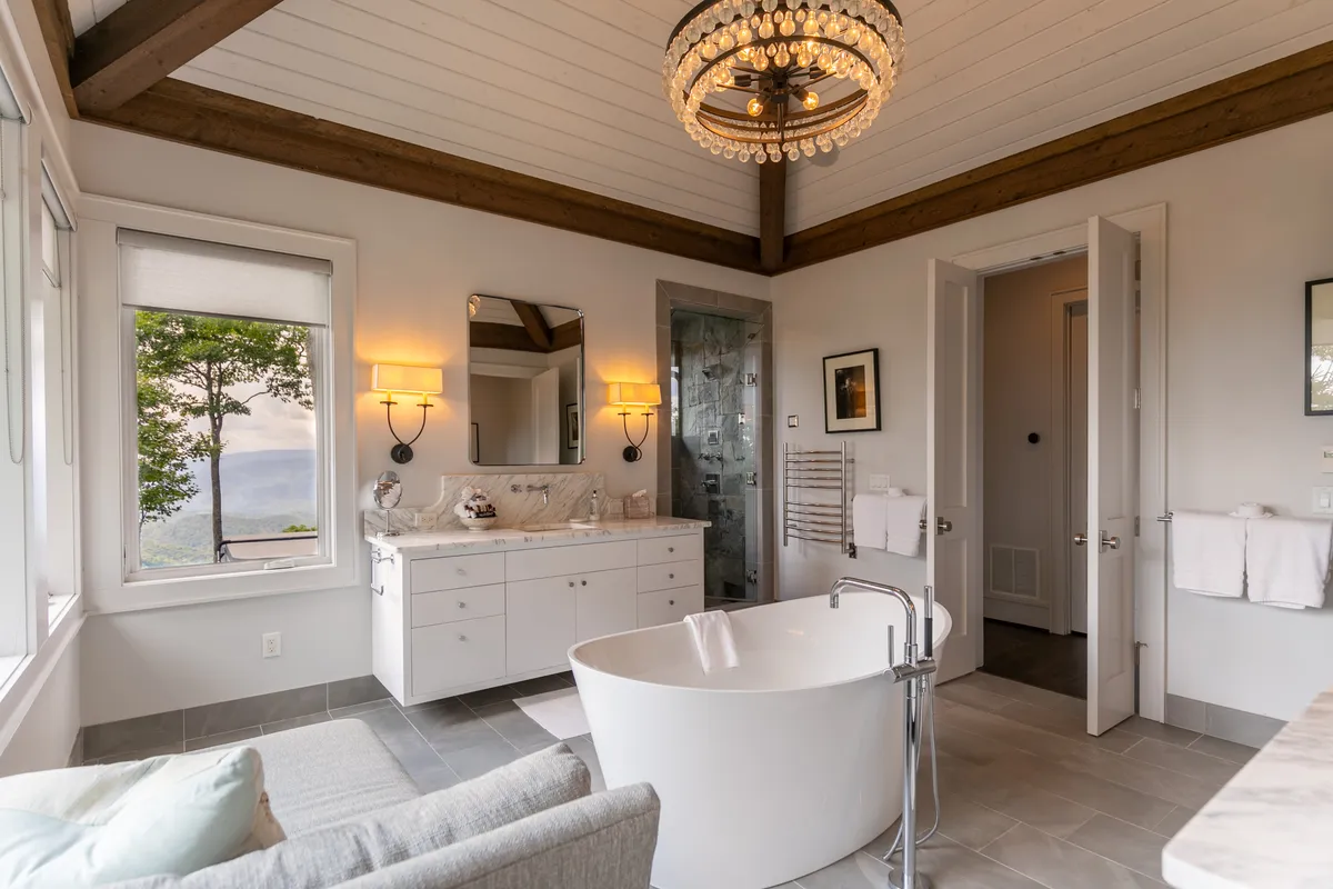 A sophisticated bathroom featuring a freestanding bathtub, elegant chandelier, marble countertop, and expansive windows framing scenic views. The luxurious space is bathed in natural light and offers a tranquil setting for indulgent self-care routines.