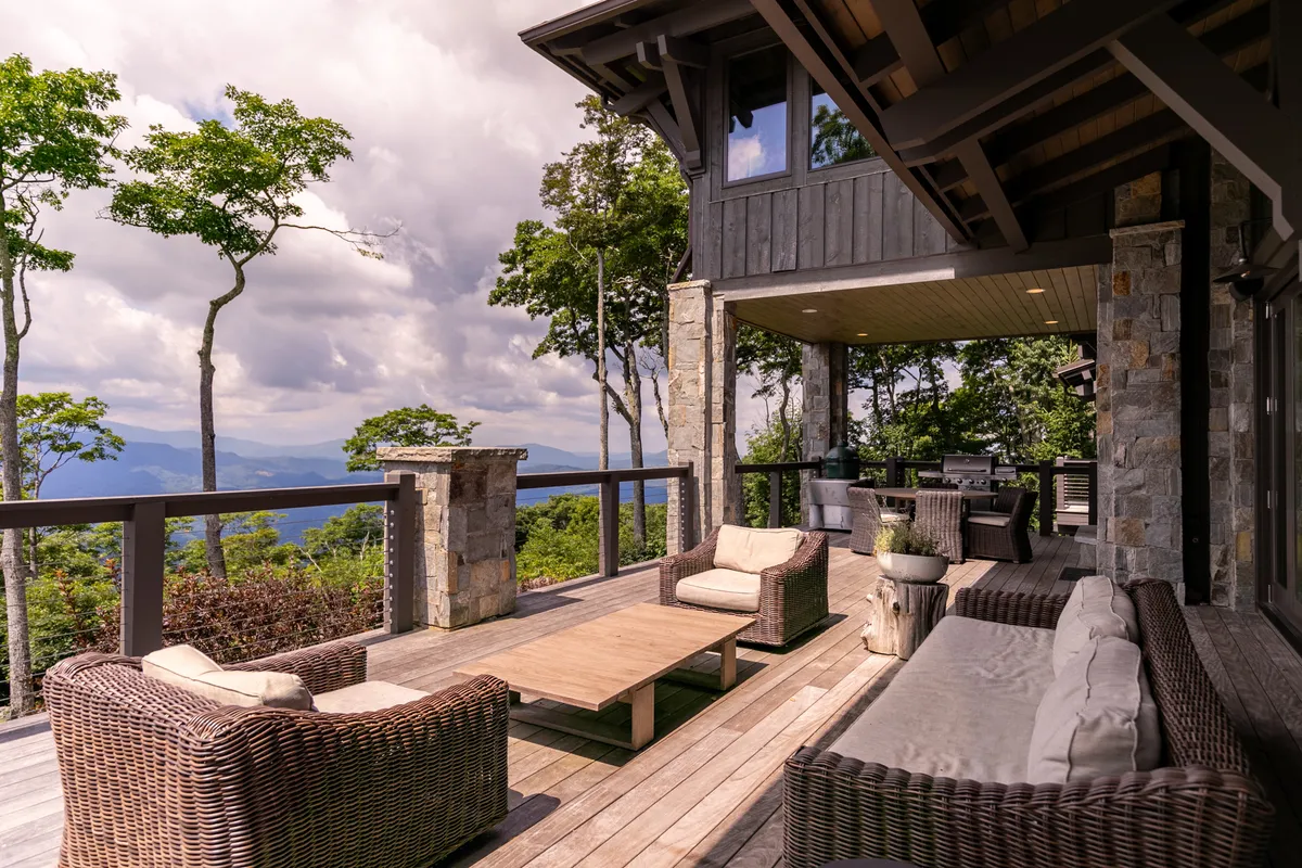 A spacious wooden deck with comfortable outdoor seating overlooks a sweeping panorama of mountain ranges. The calm mood is accentuated by the golden hues of sunset, inviting relaxation and contemplation while immersed in nature's grandeur.