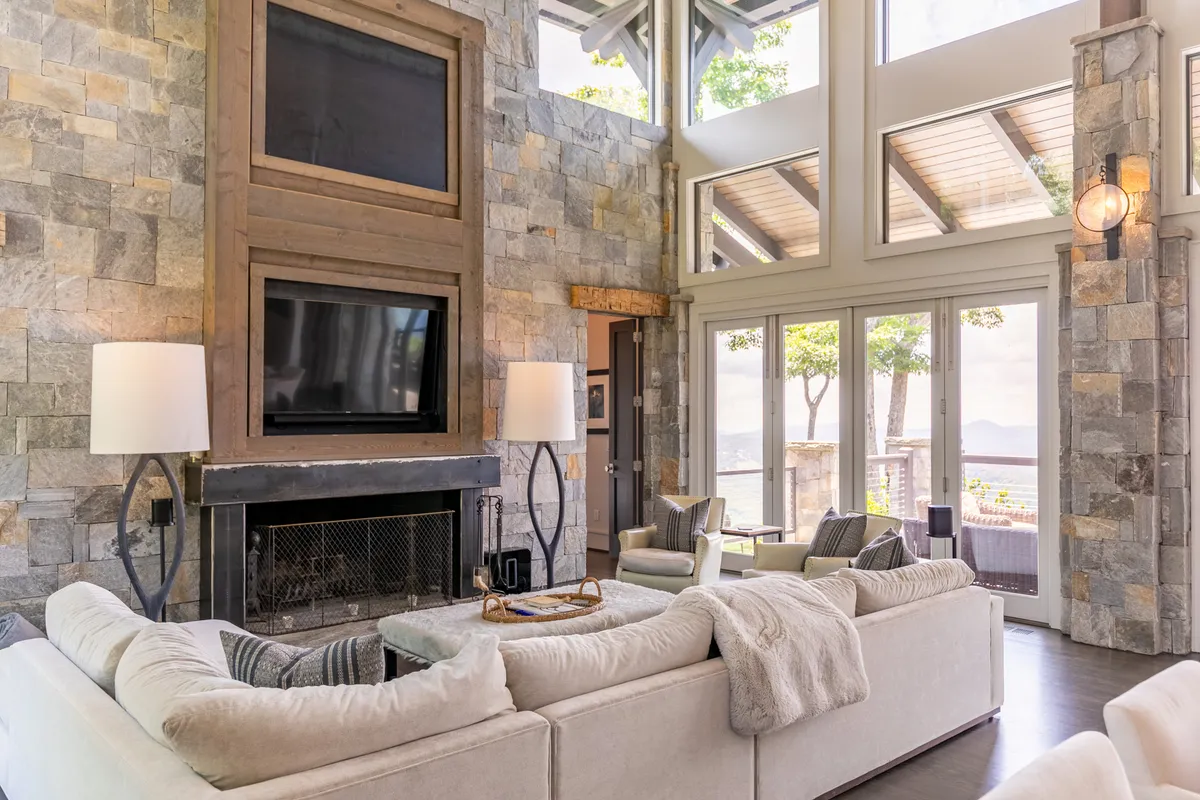 A cozy, inviting living room features a plush sofa, modern lamp, prominent stone fireplace, and large windows that allow natural light to pour in, with mountain scenery visible beyond. The space feels both contemporary and rustic, inviting relaxation and connection.