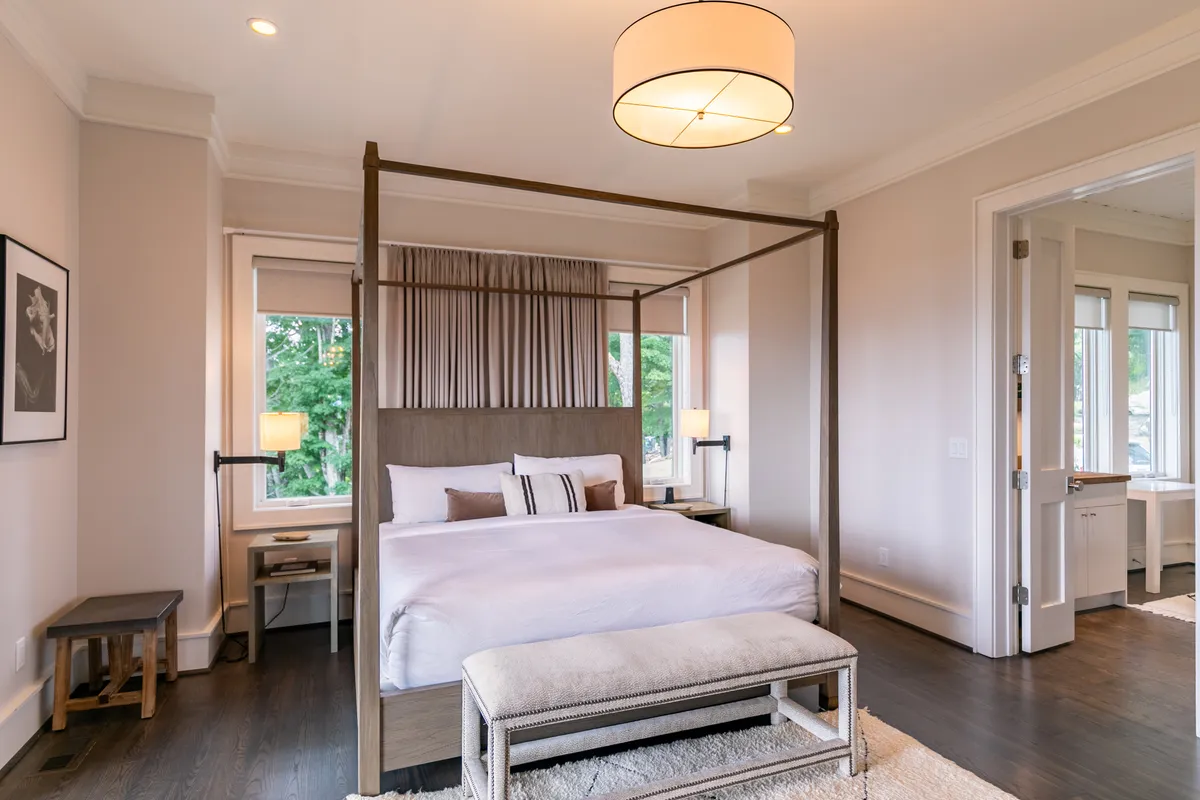 A tranquil bedroom with a four-poster bed, soft bench at the foot, large windows letting in morning sunlight, and understated artwork on the wall. The cozy furnishings and neutral tones create an inviting space that encourages relaxation and quiet moments.