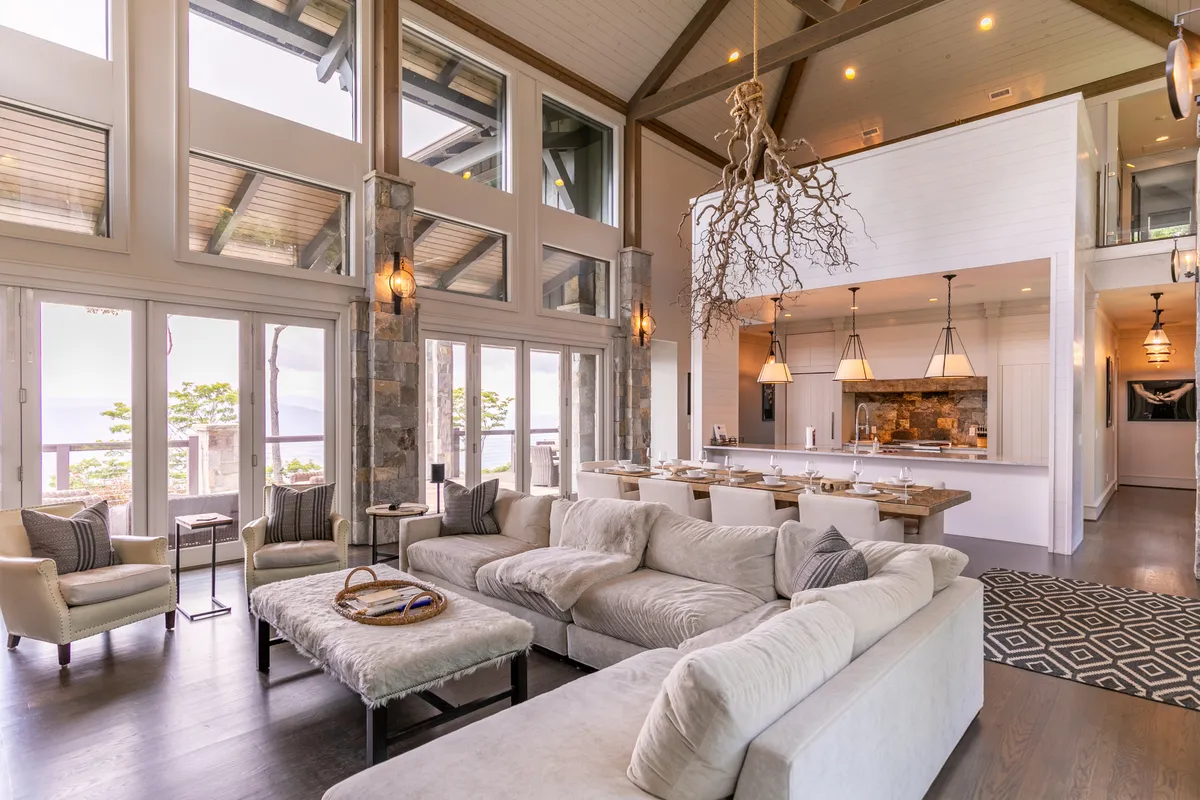 This image presents a spacious living room with a large sectional sofa, a striking stone fireplace, a modern dining area, and expansive windows that flood the space with natural light and reveal panoramic mountain vistas. There is a unique chandelier overhead, and the overall decor blends rustic elements with contemporary touches, including curated artwork.