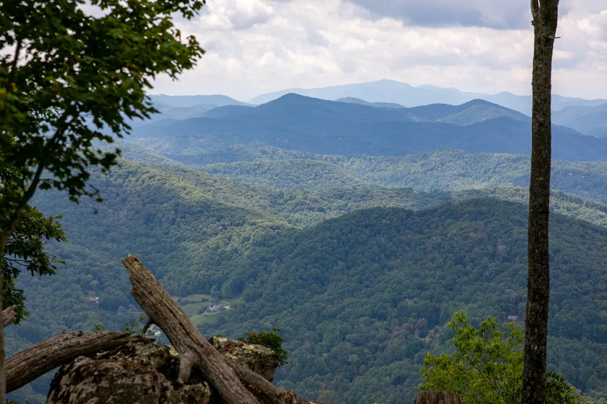 An unobstructed vista of distant, rolling mountain ranges beneath a dramatic, cloudy sky. The lush green hills and expansive wilderness evoke a deep sense of tranquility, open exploration, and connection to Appalachian scenery.