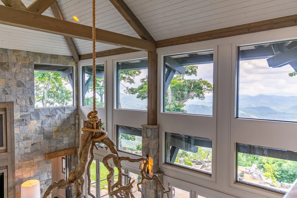 An elegant interior with large windows revealing stunning mountain views, a stone accent wall, exposed wooden beams, and a statement chandelier. The furnishings are comfortable yet sophisticated, and the architectural details highlight both craftsmanship and modern style.