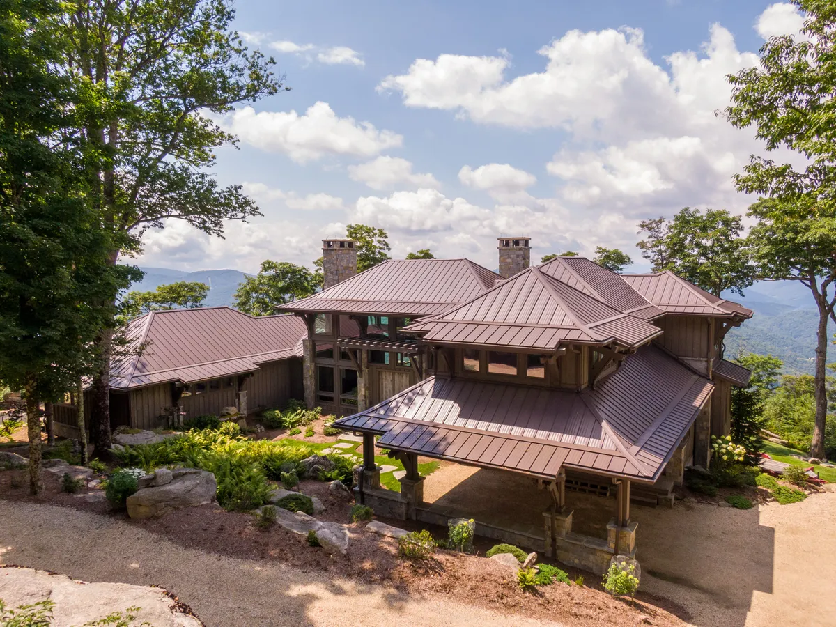 The image showcases a striking modern mountain home with stone accents and sleek metal roofing, nestled amidst lush greenery at the edge of a forest. The expansive porch offers sweeping sunset views over rolling peaks, inviting guests to relax and soak in the tranquil natural beauty surrounding the property.