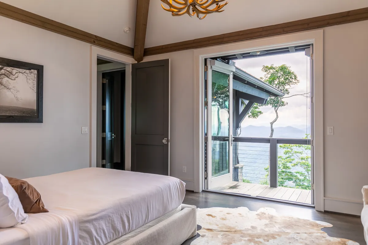 A serene bedroom featuring a plush bed dressed in soft linens, an elegant chandelier, refined decor, and an open door that leads to a private balcony. Expansive windows frame the breathtaking mountain views, filling the space with natural light and inviting the outdoors in.
